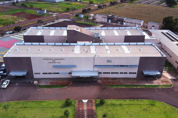 Fotografia aérea em ângulo elevado exibe o prédio do Hospital Regional de Toledo, uma estrutura moderna e horizontal de dois pavimentos com fachada em tons de bege e marrom-escuro. No centro da fachada frontal, lê-se a inscrição "HOSPITAL REGIONAL DE TOLEDO-PR" ao lado da sigla "HRT", acompanhada por marquises metálicas que cobrem três entradas. À frente do prédio há uma via asfaltada com canteiros gramados e, nos fundos e ao redor, observam-se vias de acesso, estacionamentos e áreas abertas com vegetação e campos agrícolas ao fundo sob luz do dia.
