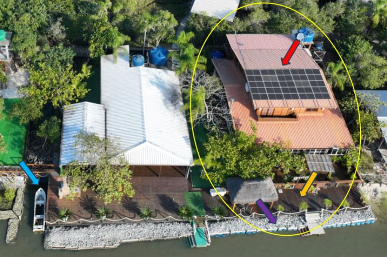 Foto aérea de duas casas à beira-rio com grandes decks de madeira. A casa da direita tem painéis solares e um gazebo, enquanto a casa da esquerda tem um pequeno barco atracado.