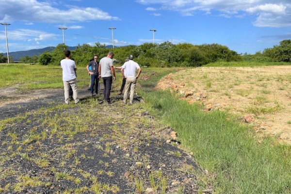 A foto mostra cinco homens do MPF em vistoria durante perícia judicial em uma área com vestígios de carvão no solo. Eles estão próximos a um canal de drenagem, que separa a parte ainda contaminada daquela onde o carvão já foi removido. Ao fundo, há vegetação, postes de luz e montanhas sob céu azul.