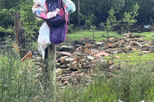 Foto colorida mostra, em primeiro plano, uma mochila meio aberta e sacolas penduradas em um poste de madeira rústico à esquerda. O poste está numa área com vegetação alta e verde. Atrás da mochila, há um monte de entulho composto por pedaços de tijolos vermelhos e destroços de construção espalhados sobre o solo. Ao fundo, há mais vegetação e algumas árvores pequenas. O dia está nublado.