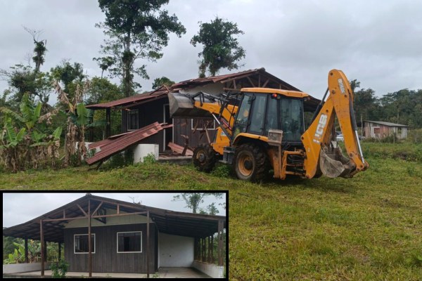 Imagem de uma retroescavadeira demolindo uma casa de madeira. O veículo levanta parte do telhado já arrancado. No canto inferior esquerdo, em destaque, aparece uma foto da mesma casa antes da demolição, com paredes de madeira e varanda coberta.