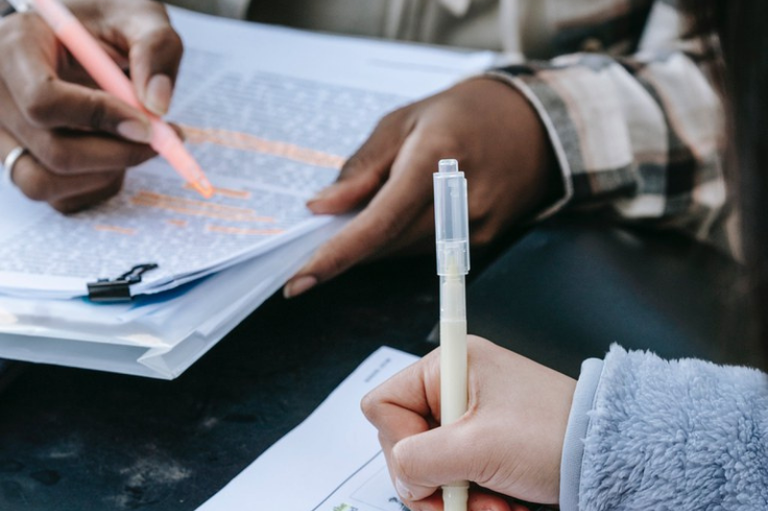 a imagem em plano detalhado mostra as mãos de duas pessoas sobre uma superfície escura, em uma atividade de análise de documentos. No canto superior esquerdo, as mãos de uma pessoa negra seguram uma caneta marca-texto de cor laranja, que está sendo utilizada para destacar trechos de um texto em uma folha branca presa por um clipe metálico preto. Próximo ao centro, a mão de outra pessoa, de pele clara e vestindo um casaco de pelúcia cinza, segura uma caneta branca com tampa transparente, posicionada como se estivesse pronta para escrever em outra folha de papel. A iluminação é natural e clara, focando na interação entre as mãos e os papéis, enquanto o fundo permanece desfocado.