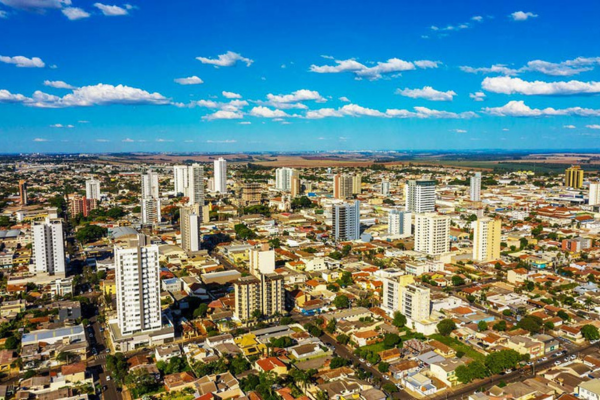Fotografia aérea em ângulo panorâmico mostra o centro urbano de Arapongas em um dia ensolarado sob céu azul com pequenas nuvens brancas. Em primeiro plano e na parte central, destacam-se diversos edifícios residenciais e comerciais de médio e grande porte, distribuídos entre uma densa malha de casas térreas e sobrados. Árvores verdes pontuam as ruas e avenidas ao longo do traçado urbano, enquanto ao fundo sobressaem grandes áreas planas de campos agrícolas de tom marrom e a linha do horizonte.