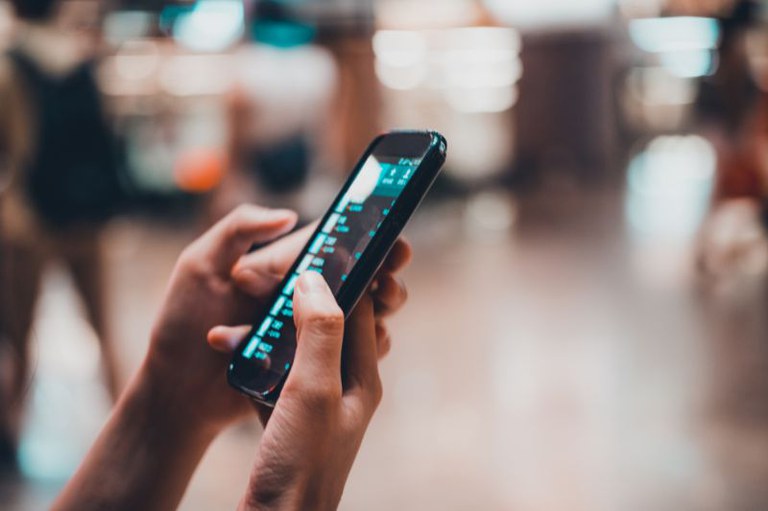 Mãos de uma pessoa segurando e manuseando um smartphone, em ambiente interno com fundo desfocado.
