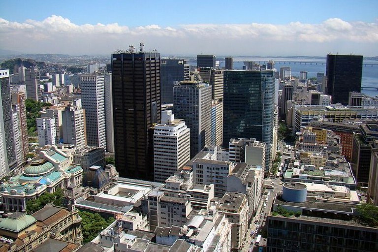 Uma vista aérea exibe o denso centro urbano do Rio de Janeiro, com arranha-céus modernos contrastando com edifícios históricos de arquitetura clássica.