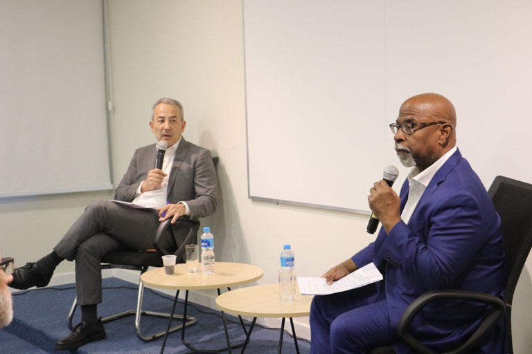 Dois homens estão sentados em cadeiras de escritório, conversando com microfones em mãos, em um ambiente interno de paredes claras e carpete azul. O homem à esquerda veste terno cinza, camisa branca e está com as pernas cruzadas, segurando um bloco de anotações. O homem à direita, identificado como o professor Kendall Thomas, usa terno azul e camisa branca, e segura folhas de papel. Entre eles há duas mesas redondas baixas com garrafas e copos de água, além de um copo descartável com café. Ao fundo, há um quadro branco e uma persiana cinza.