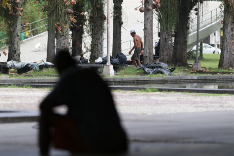 foto de um morador de rua andando andando embaixo de árvores, em primeiro plano uma pessoa sentada de frente para a cena
