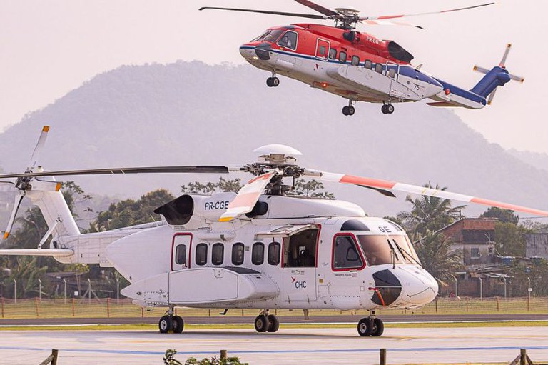 Foto mostra um helicóptero branco no chão e outro da cor vermelha decolando