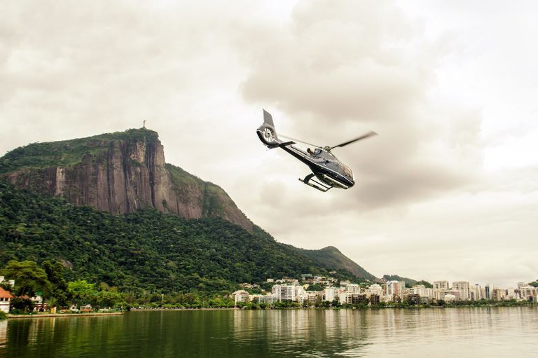 Um helicóptero preto em voo sobre uma lagoa. No fundo, a montanha Dois Irmãos com uma cruz no topo e a paisagem urbana sob um céu nublado com tom sépia.