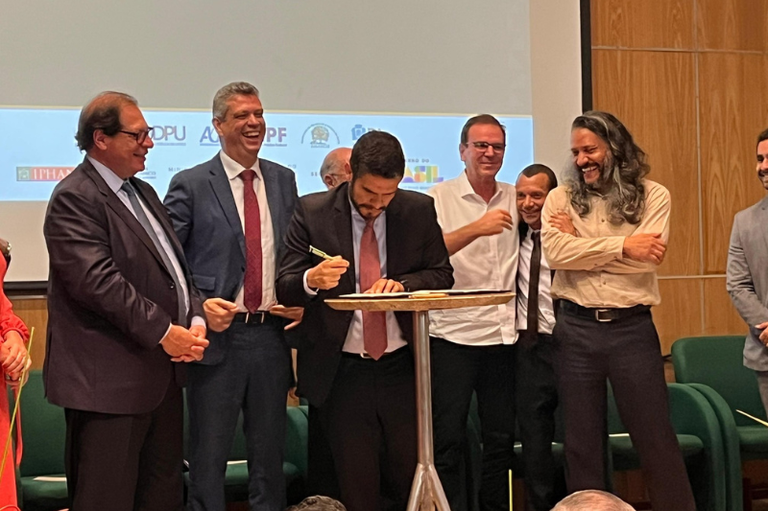 Um grupo de sete homens, a maioria em trajes formais (ternos), está reunido em uma cerimônia de assinatura. O homem ao centro assina um documento em uma pequena mesa, enquanto os demais o observam, muitos deles sorrindo ou rindo. Um logo grande com as letras DPU (Defensoria Pública da União) é visível no fundo.