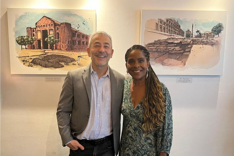 Duas pessoas posam lado a lado, sorrindo para a câmera, em um ambiente interno com paredes claras. À esquerda, um homem de cabelos grisalhos veste blazer cinza, camisa clara e calça escura, com uma das mãos no bolso. À direita, uma mulher de cabelos longos trançados veste um vestido estampado em tons de verde e brincos grandes. Ao fundo, há duas obras de arte emolduradas com ilustrações de construções e paisagens urbanas.