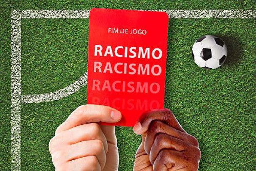 Foto mostra um gramado ao fundo. Em primeiro plano uma pessoa com a mão branca e uma com a mão negra mostram um cartão vermelho. Nele está escrito Racismo Fim de Jogo