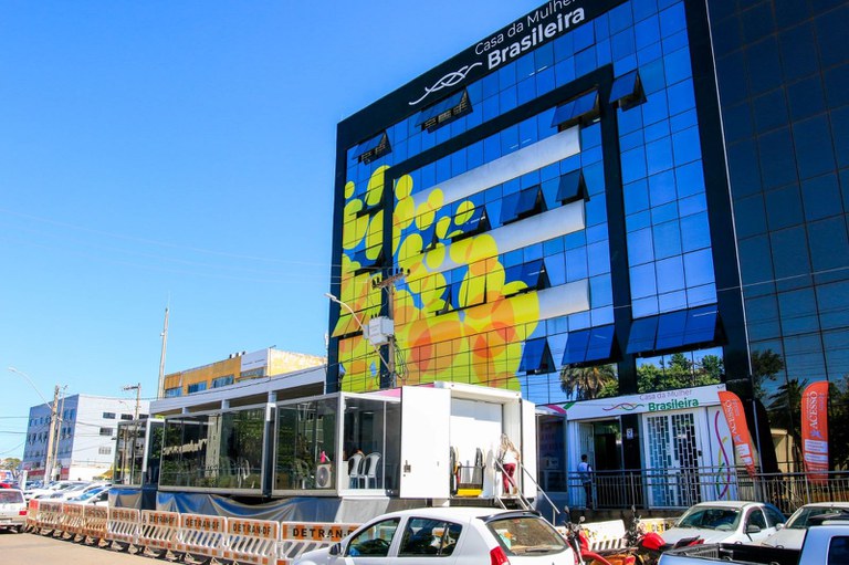 Fotografia da Casa da Mulher Brasileira de Ceilândia, no Distrito federal, na qual há a fachada de vidro do prédio, na cor azul com adesivos amarelos, e uma rua em frente, com um container e alguns carros.