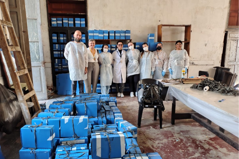Foto de um grupo de oito profissionais em uma sala de arquivo com paredes desgastadas. Eles estão posicionados atrás de dezenas de caixas de arquivo azuis empilhadas no chão e em estantes ao fundo. A maioria veste aventais descartáveis brancos e máscaras de proteção. À direita, uma mesa de trabalho contém documentos e materiais de conservação; à esquerda, uma escada de madeira encostada na parede.