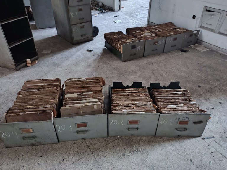 A imagem mostra gavetas de arquivo de metal cinza organizadas no chão de uma sala abandonada e desgastada. As gavetas estão numeradas à mão e repletas de pastas suspensas e documentos antigos de papel pardo.