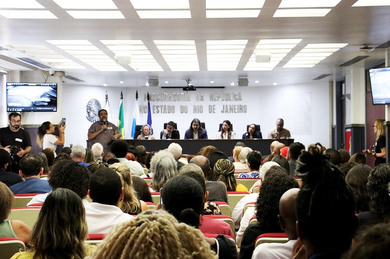 A imagem mostra uma audiência pública em um auditório cheio de pessoas. No centro, há uma mesa com vários palestrantes sentados, enquanto um homem em pé fala ao microfone. O ambiente tem bandeiras, telões laterais e um letreiro ao fundo indicando que o evento ocorre na Procuradoria da República no Estado do Rio de Janeiro.