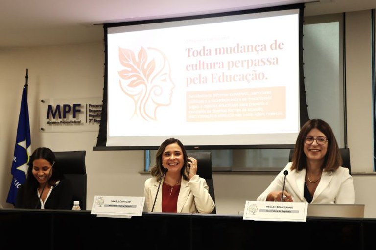 Foto mostra três mulheres com roupas formais sentadas à mesa do evento com microfones e placas com os nomes e cargos, por trás um telão com a frase toda mudança de cultura perpassa pela educação