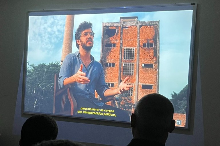 Fotografia de uma projeção numa sala escura. A imagem projetada mostra um homem jovem, de cabelo escuro, barba e óculos, a usar uma camisa azul, sentado numa cadeira de madeira e a gesticular com as mãos abertas enquanto fala. Ao fundo da cena projetada, vê-se a estrutura em tijolo e cimento de um edifício antigo e em ruínas, além de uma chaminé alta do lado esquerdo. Na parte inferior da projeção, surgem legendas em amarelo com o texto: "para incinerar os corpos dos desaparecidos políticos,".