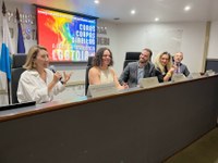 Cinco pessoas estão sentadas lado a lado atrás de uma longa bancada em um auditório. Elas participam de um evento, aparentando estar em uma mesa de debate. Ao fundo, um telão exibe uma imagem colorida com as palavras "Corpos, Cores, Direitos – Exposição LGBTQIA+". À esquerda da imagem, há bandeiras posicionadas verticalmente. As pessoas estão bem iluminadas, sorrindo ou falando ao microfone, e o ambiente transmite um clima de acolhimento e celebração da diversidade.