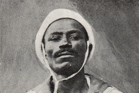 Fotografia antiga em preto e branco mostra o rosto de João Cândido, líder da Revolta da Chibata, um homem de pele negra, bigode, que olha em frente. Ele usa o chapéu e a roupa branca da marinha.