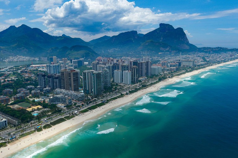 Esta deslumbrante vista aérea captura a orla da Barra da Tijuca, no Rio de Janeiro, revelando o contraste marcante entre o azul-turquesa do Oceano Atlântico e a densa urbanização à beira-mar. A extensa faixa de areia clara é banhada por ondas suaves e margeada por uma avenida movimentada, onde se alinham modernos arranha-céus residenciais e comerciais que definem o horizonte do bairro. Ao fundo, a paisagem é emoldurada pelas imponentes formações rochosas e montanhas cobertas por vegetação nativa, como a Pedra da Gávea, sob um céu dinâmico com nuvens brancas volumosas que conferem profundidade e grandiosidade ao cenário litorâneo.