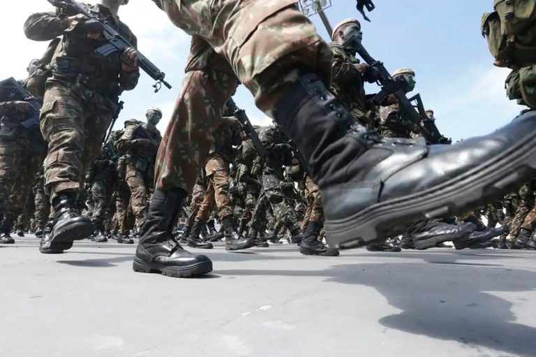 foto mostra militares do exercito com uniforme camuflado e armas nas mãos, marchando
