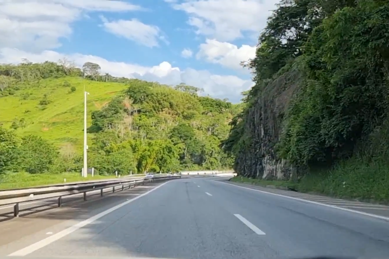 Fotografia em plano geral da rodovia pavimentada BR-040, com pista dupla, sinalização horizontal em branco e defensas metálicas à esquerda. Ao lado direito, sobressai-se um paredão rochoso elevado coberto por vegetação densa e árvores, enquanto à esquerda estendem-se morros verdes sob um céu ensolarado com poucas nuvens brancas.