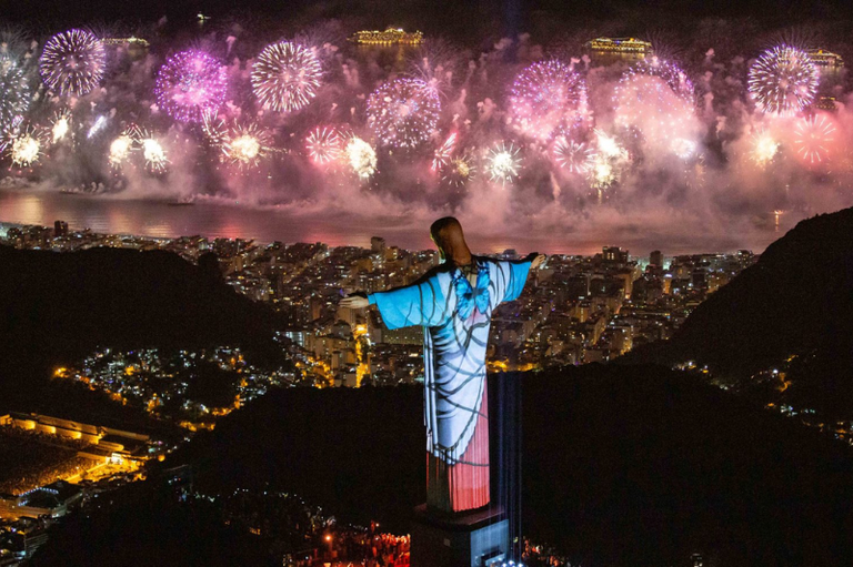 Foto noturna de queima de fogos de artifício na praia de Copacabana; em primeiro plano, vê-se a estátua do Cristo Redentor de costas