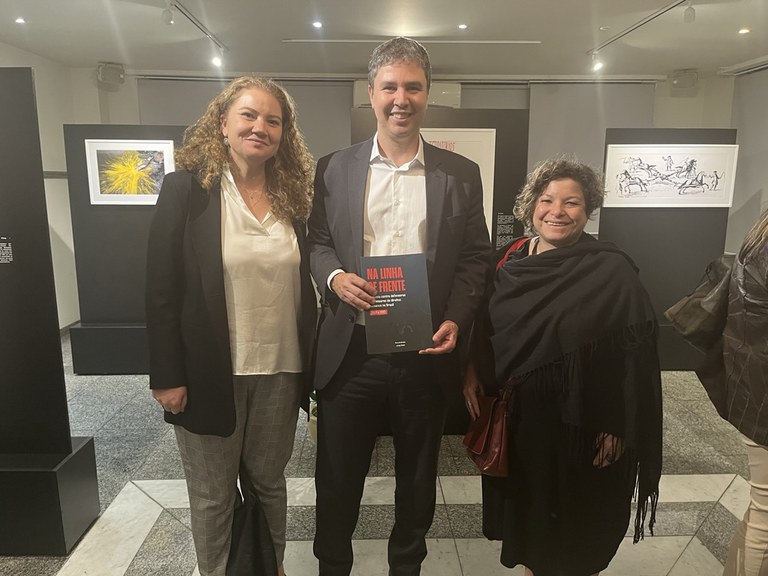 A imagem mostra três pessoas posando lado a lado em um espaço expositivo. À esquerda está uma mulher de cabelos cacheados, loiros e soltos, vestida com blazer preto, blusa branca e calça xadrez em tons de cinza. Ao centro, um homem de terno escuro e camisa branca segura com as duas mãos um livro ou catálogo com a capa preta e título em vermelho que diz “Na Linha de Frente”. À direita, outra mulher, de cabelos curtos e cacheados, veste um xale preto sobre a roupa e carrega uma bolsa de ombro vermelha. Ao fundo, há paredes pretas com quadros emoldurados exibindo obras de arte. O chão é de mármore e granito em tons de cinza e branco.