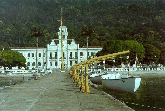 Fotografia de um prédio antigo, em estilo colonial ao fundo, cercado por floresta, e, em primeiro plano, um cais, com barcos na direita.