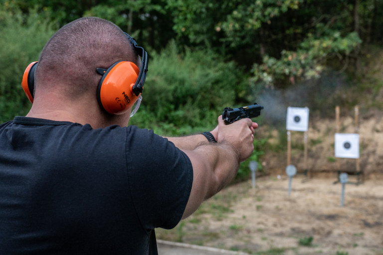 Foto mostra homem com camisa preta e abafador de ruídos alaranjado nos ouvidos, de costas para o espectador, com uma pistola nas mãos apontada para um alvo, em área de mata