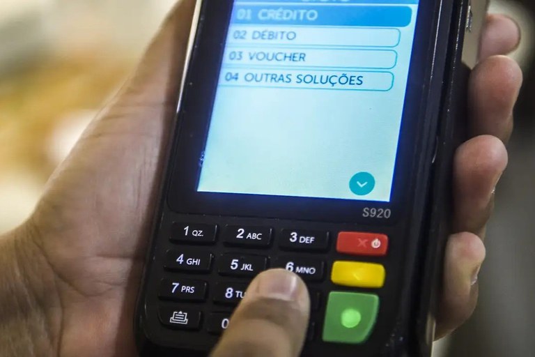 A imagem mostra um close-up das mãos de uma pessoa operando uma maquininha de cartão de crédito preta (modelo S920). A tela colorida do dispositivo exibe um menu em português com as opções de pagamento: 01 CRÉDITO, 02 DÉBITO, 03 VOUCHER e 04 OUTRAS SOLUÇÕES, estando a primeira opção destacada em azul. O dedo indicador do usuário aparece pressionando a tecla numérica 6 no teclado físico, que possui botões de borracha pretos com números e letras brancas, além das teclas coloridas padrão (vermelha para cancelar, amarela para corrigir e verde para confirmar). O fundo está desfocado, mantendo o foco total na transação financeira em andamento.