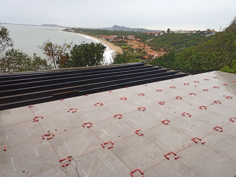 Foto em plano aberto das obras do mirante. A imagem mostra um piso sendo instalado em um terraço com vista para o mar. Em primeiro plano, o piso de cor cinza claro com veios está assentado sobre o contrapiso. Peças plásticas vermelhas, usadas como espaçadores e niveladores, estão distribuídas sobre as juntas das placas em formação de grade. À esquerda, uma estrutura preta em faixas horizontais sugere uma área de deck em construção.