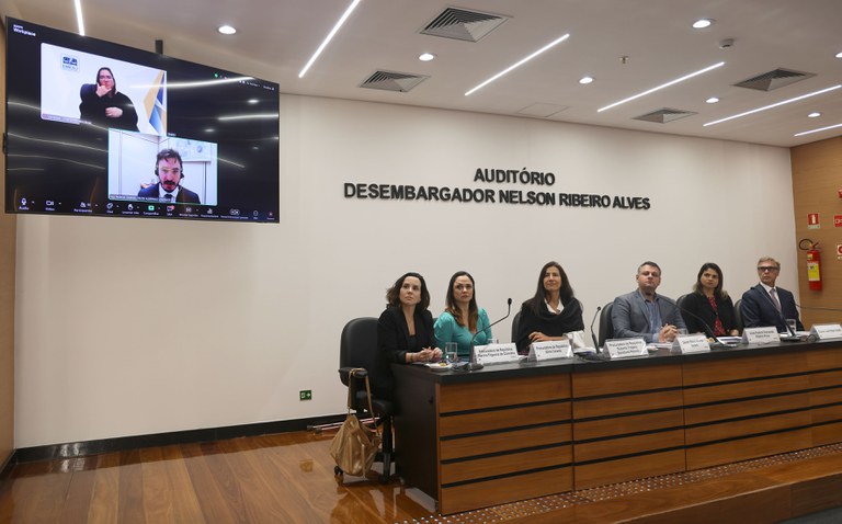 Fotografia colorida tirada no Auditório Desembargador Nelson Ribeiro Alves, com sete pessoas participando de um evento institucional. Seis pessoas estão sentadas à mesa principal, voltadas para o público, e uma participa por videoconferência, exibida em um telão à esquerda da imagem. A parede ao fundo é clara, com o nome do auditório em letras pretas. No telão, aparecem dois quadros: o superior mostra uma intérprete de Libras e o inferior mostra um homem de terno participando remotamente. À mesa presencial, há três mulheres e três homens, todos com identificação em frente. O ambiente é bem iluminado, com piso de madeira e teto com luzes embutidas. A expressão dos participantes é séria e atenta, sugerindo uma ocasião formal, como uma audiência pública ou seminário jurídico.