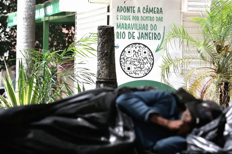 Foto mostra, em primeiro plano desfocado, homem deitado em área de rua, coberto por sacos de lixo pretos; ao fundo, vê-se uma estrutura de quiosque com a inscrição "Aponte a câmera e fique por dentro das maravilhas do Rio de Janeiro"