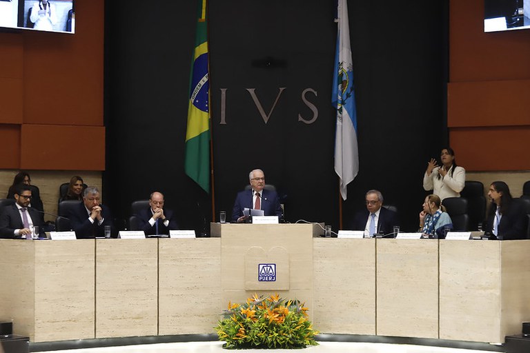 Fotografia horizontal em plano médio de uma solenidade judiciária. Sete pessoas ocupam uma bancada de mármore claro; ao centro, um homem de terno azul discursa com papéis em mãos. À frente da bancada, há o logotipo do PJERJ e um arranjo de flores tropicais. Ao fundo, painéis de madeira laranja ladeiam uma parede preta com a sigla "IVS" em prata, posicionada entre as bandeiras do Brasil e do Estado do Rio de Janeiro. À direita, uma intérprete de Libras faz sinais. No topo, monitores exibem a transmissão do evento.