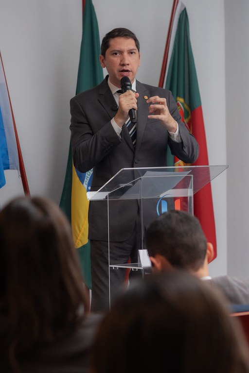 Procurador da República Luís Cláudio Consentino está em pé atrás de um púlpito de acrílico transparente, segurando um microfone com a mão direita enquanto gesticula com a esquerda durante sua fala em uma audiência pública. Ele veste terno cinza, camisa branca e gravata listrada em tons de preto, cinza e branco. Ao fundo, aparecem as bandeiras do Brasil e do Rio Grande do Sul, além de outra bandeira parcialmente visível. Em primeiro plano, desfocadas, estão as cabeças de pessoas da plateia, reforçando o contexto de um evento com público presente. A expressão do procurador é séria e concentrada, transmitindo o momento de exposição e debate do tema.