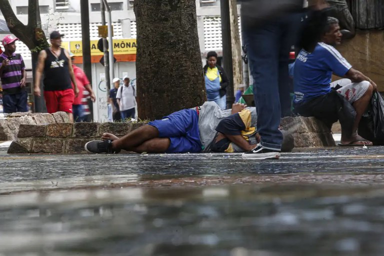 A imagem captura uma cena urbana em local público, onde um homem está deitado no chão de paralelepípedos, vestindo bermuda azul e camiseta nas cores cinza e amarela. Ao fundo e nas proximidades, outras pessoas circulam ou estão sentadas em muretas de pedra sob a sombra de uma árvore, enquanto fachadas comerciais aparecem desfocadas no cenário. O chão apresenta aspecto úmido, refletindo discretamente a iluminação do ambiente.