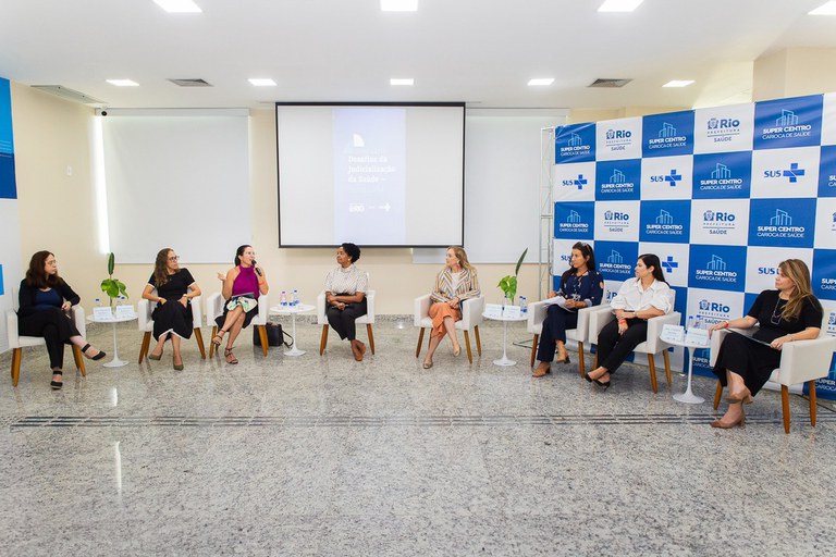 Foto mostra um grupo de mulheres debatendo durante uma seminário. Uma delas está falando ao microfone, enquanto as demais escutam. Ao fundo, um enorme banner de lona mostra logomarca do Sistema Único de Saúde (SUS)e da prefeitura do Rio.
