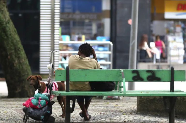 Foto mostra uma moradora de rua sentada em um banco de praça