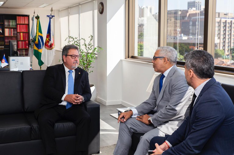 Fotografia de uma reunião realizada em um gabinete institucional. À esquerda, está sentado em um sofá preto o procurador-geral de Justiça do Rio de Janeiro, Antonio José Campos Moreira, vestindo terno preto, camisa branca e gravata azul clara. À direita da imagem, estão o procurador da República Eduardo Benones, de terno cinza claro, e outro homem de terno escuro, ambos sentados em frente ao procurador-geral. Os três conversam em ambiente iluminado por luz natural, com janelas amplas ao fundo. Ao lado de uma estante com livros, aparecem as bandeiras do Brasil, do Estado do Rio de Janeiro e do Ministério Público. A reunião teve como objetivo alinhar estratégias de cooperação entre o MPRJ e o MPF no controle externo da atividade policial.