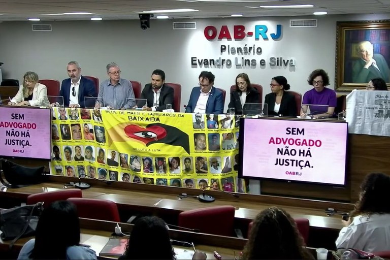 MPF e parceiros estabelecem agenda de enfrentamento dos desaparecimentos Fotografia de uma reunião no auditório da sede da OAB/RJ, com autoridades sentadas à mesa e pessoas na platéia. Em duas telas na mesa do evento há a mensagem "sem advogado não há justiça"