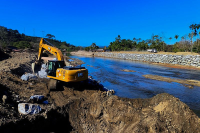 Fotografia de uma escavadeira amarela (marca CAT) operando às margens do Rio Bracuí, em Angra dos Reis (RJ), em um cenário de obras de intervenção. O equipamento movimenta grandes pedras junto a um barranco de terra, com o leito do rio parcialmente visível ao lado direito. O céu está limpo e azul, e ao fundo vê-se vegetação densa, algumas palmeiras e outra máquina ao longe. As margens do rio estão reforçadas com enrocamento de pedras. A cena ocorre em um ambiente rural e ensolarado.