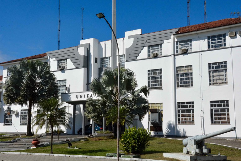 Fotografia em ângulo aberto da fachada principal da Universidade da Força Aérea (UNIFA), um prédio branco de múltiplos andares com janelas em fita e arquitetura moderna. O nome "UNIVERSIDADE DA FORÇA AÉREA" e o símbolo da FAB (estrela com asas) estão em destaque no topo. Em primeiro plano, uma área de gramado bem cuidado e, ao fundo, um céu azul claro, reforçando a imponência e o contexto militar-aeronáutico da instituição.