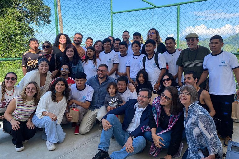 A imagem mostra um grupo diverso de pessoas reunidas ao ar livre. Ao fundo, há uma cerca de tela verde com paisagem montanhosa visível. Algumas pessoas usam camisetas brancas com logotipo escolar.
