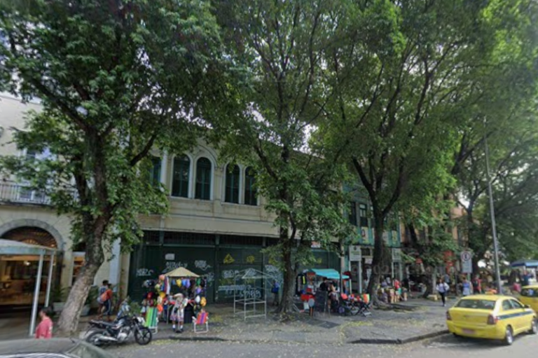 Fotografia ao nível da rua de prédios históricos na Rua do Catete, Rio de Janeiro. A calçada está arborizada com árvores altas que cobrem parcialmente as fachadas de dois andares com janelas em arco. No térreo, há portões de metal com pichações e pequenas barracas de comércio popular. Um táxi amarelo circula à direita, e pedestres caminham ao fundo.