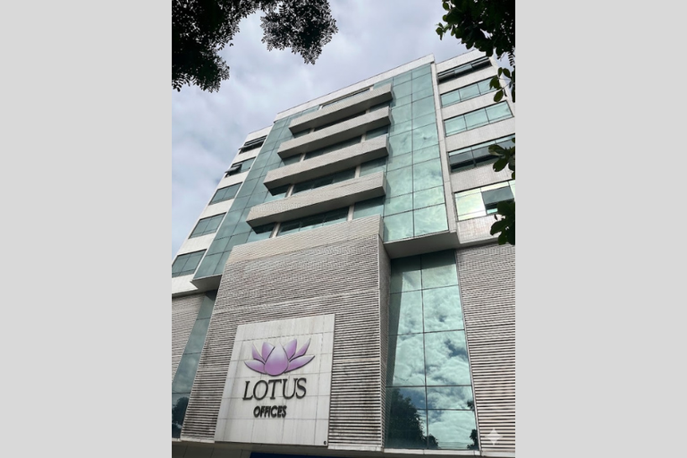 Fotografia em ângulo inferior do edifício "Lotus Offices". A fachada moderna combina pastilhas brancas, extensas superfícies de vidro espelhado esverdeado que refletem o céu nublado e painéis com frisos horizontais. Em destaque na parte inferior central, um painel branco exibe o logotipo: uma flor de lótus estilizada em tons de lilás sobre o nome "LOTUS OFFICES" em letras pretas.
