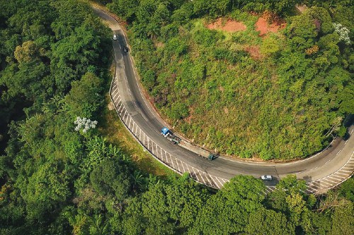 Fotografia aérea mostra um trecho sinuoso da BR-040 cercado por vegetação densa de Mata Atlântica. A pista faz uma curva acentuada em formato de ferradura, contornando uma encosta coberta por áreas verdes e trechos de terra exposta. Alguns veículos trafegam pela rodovia, incluindo um caminhão azul próximo ao centro da imagem e carros distribuídos ao longo da curva. A cena destaca o relevo montanhoso e a integração da estrada com a paisagem natural.