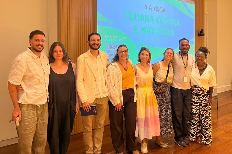 Sete pessoas estão perfiladas e a sorrir num auditório, em frente a um ecrã de projeção.   Ao fundo, o ecrã exibe a mensagem "9 A 12/10 SEMANA CRIANÇA E NATUREZA" em letras verdes sobre um fundo com tons de azul e verde. Da esquerda para a direita: Um homem vestindo camisola clara de mangas curtas e calças beges. Uma mulher de cabelo comprido vestindo macacão preto. Um homem de óculos e barba, com blazer claro e calças beges, segurando um pequeno caderno. Uma mulher de óculos, com camisa branca aberta sobre uma blusa amarela e calças escuras. Uma mulher com fato/vestido de saia amarela às cores e topo claro. Uma mulher com top claro e saia comprida estampada. Um homem de t-shirt branca e crachá ao pescoço, abraçado a uma mulher com blusa branca e calças largas com padrões pretos e brancos.