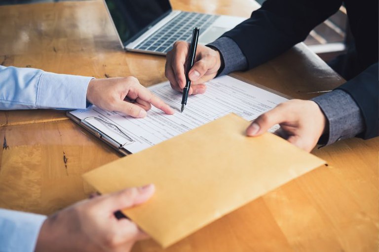 Foto mostra uma pessoa assinando um contrato enquanto outra entrega um envelope